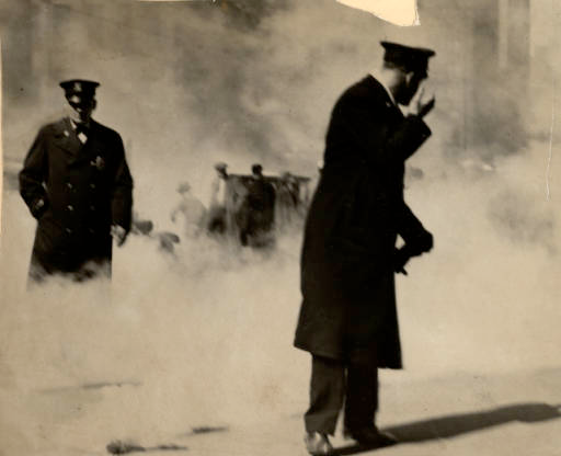 Two police stand in the foreground of the photo amidst a large cloud of smoke, strikers can be seen standing in the background.