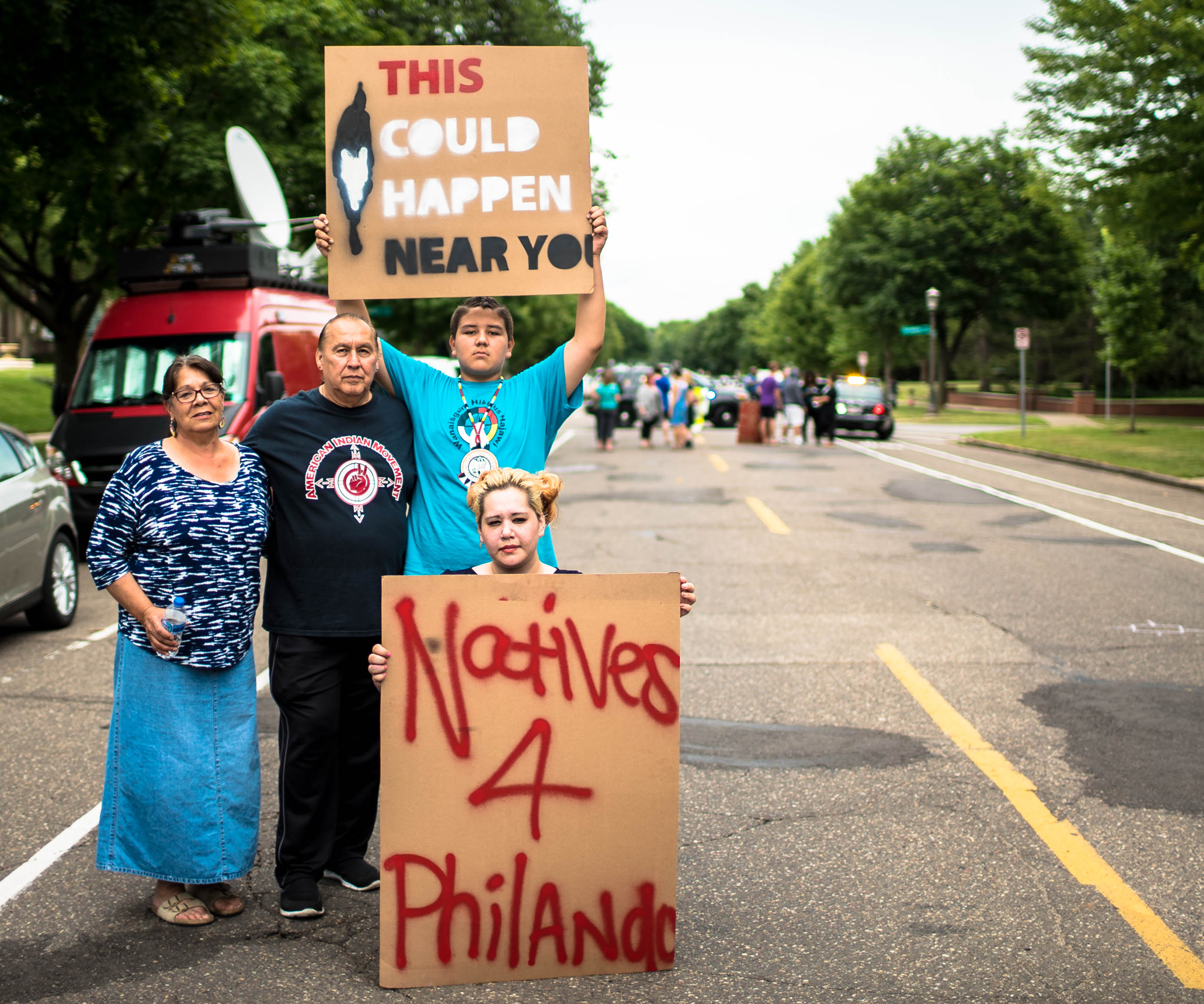 A group of 4 Native American folks stand on the side of a blockaded road. They hold signs reading, "Natives 4 Philando," and "This could happen near you."