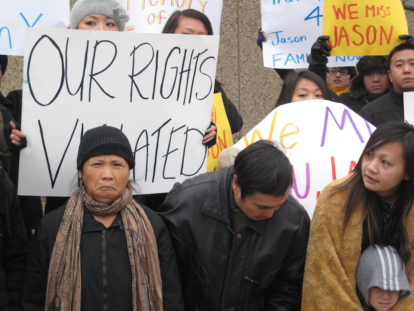 Jason Yang's parents stand centered in the photo, his mother looks upset and father looking down, behind them someone holds a sign that reads "our rights violated" another sign further back reads "we miss Jason"