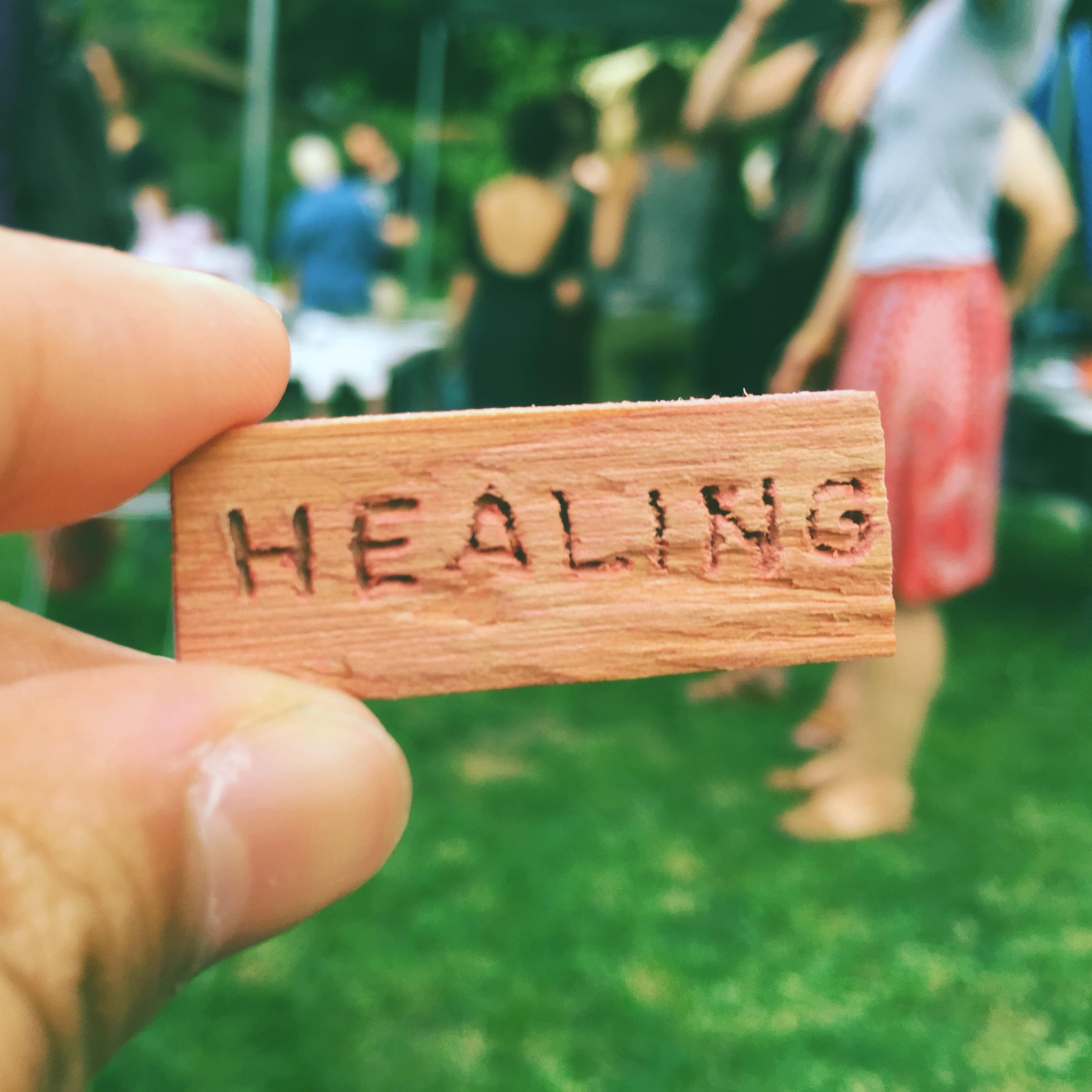 A hand holds a small, wooden block with the word "healing" carved into it. The out-of-focus background features people talking outside in a greenspace.