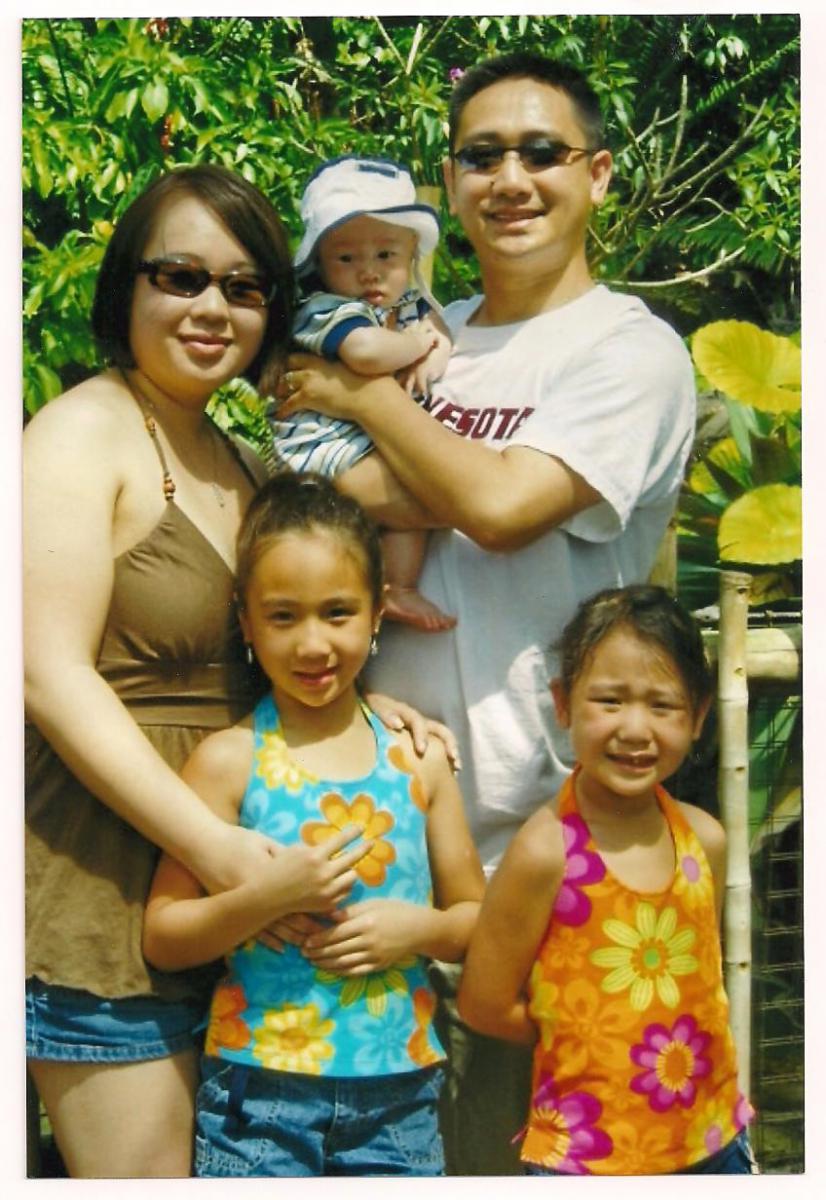 Jason Yang and family in the sun, Jason holds his baby, his wife and two daughters stand next to him. It appears to be a sunny day and they stand in front of lots of greenery.