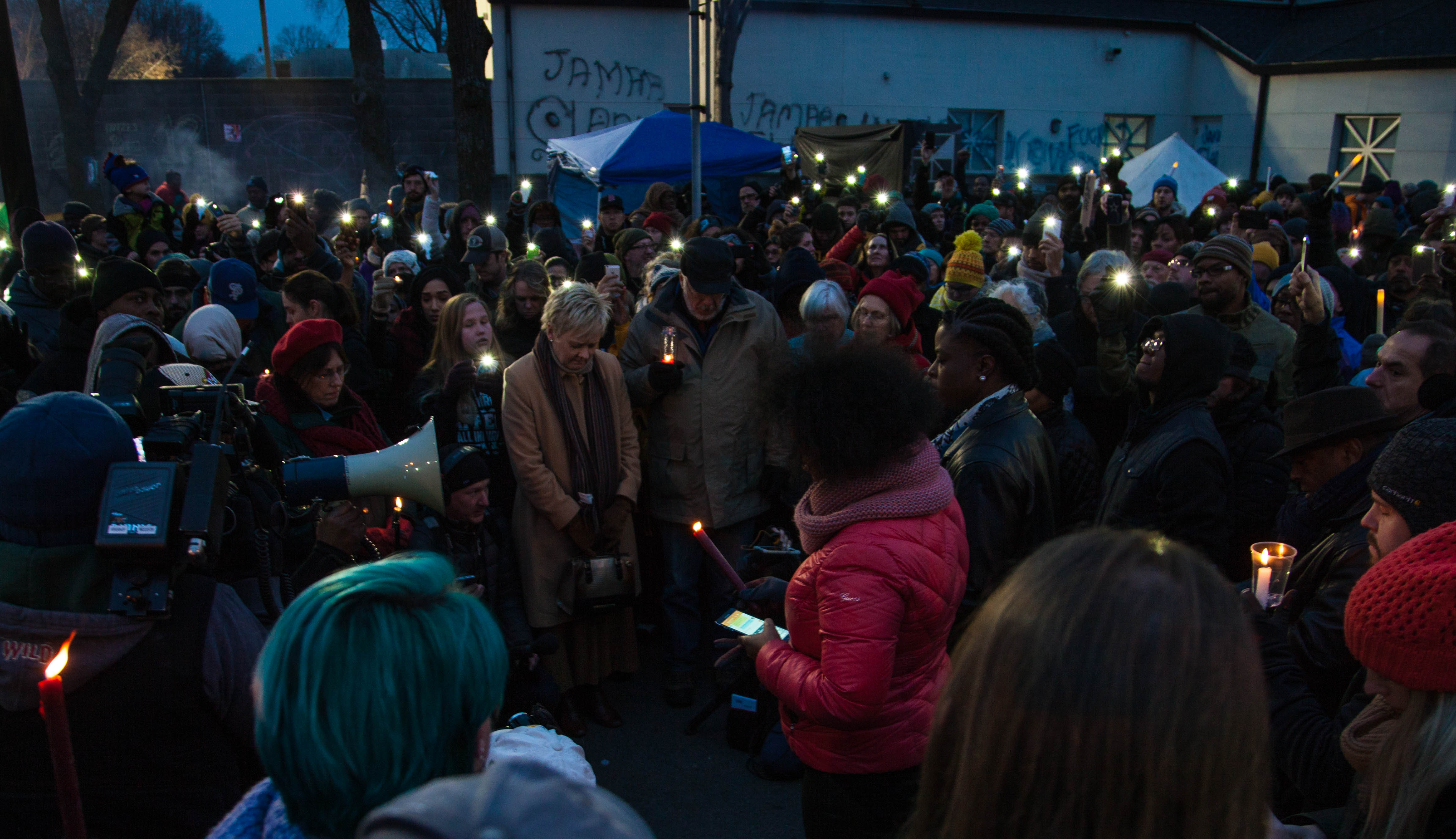 A large group of folks gathered at a candlelight vigil for Jamar Clark. It is night-time, and people have solemnly have their heads down.