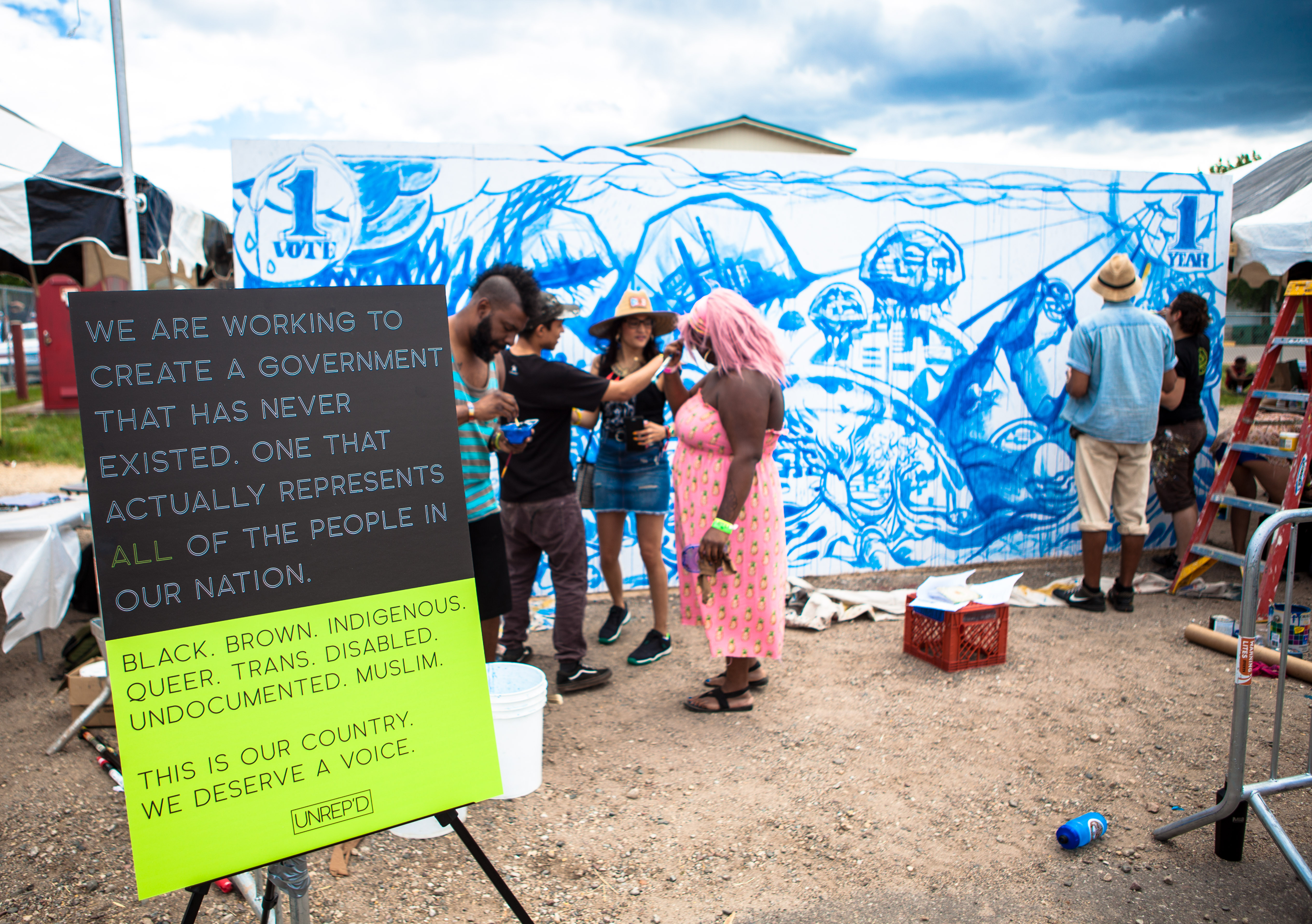 A group of artists stand in front of the beginnings of a mural on a cloudy day. In the foreground, a sign reads, "We are working to create a government that has never existed. One that actually represents all of the people in our nation. Black, brown, Indigenous, queer, trans, disabled, undocumented, Muslim. This is a country. We deserve a voice. UNREPD.