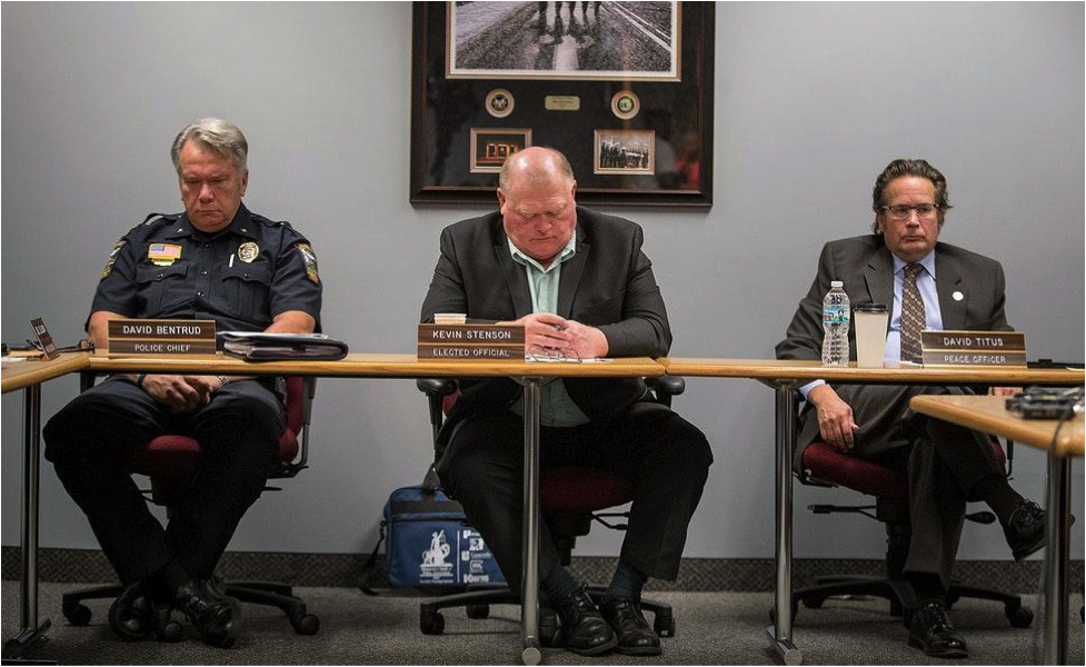Three sleepy looking white presenting men, members of the Minnesota Peace Officer Standards and Training Board sit at a wooden table.