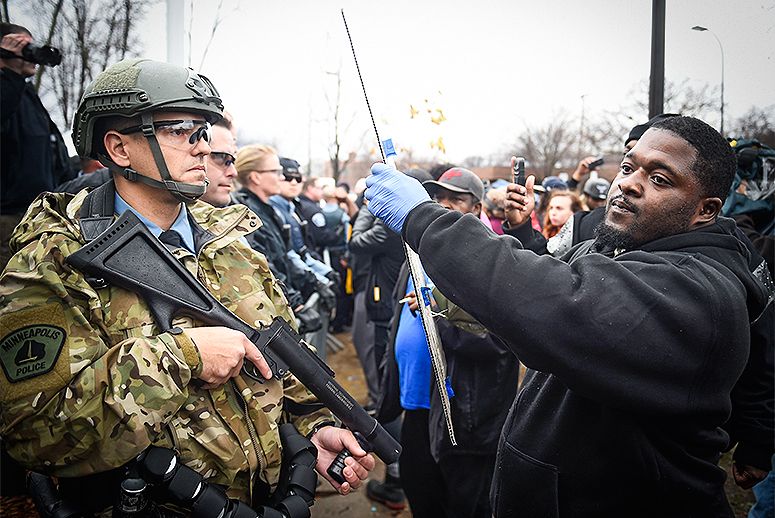 A black protester holds a sign high in the face of a white police officer, the police officer is wearing camo uniform and holding a less lethal weapon.