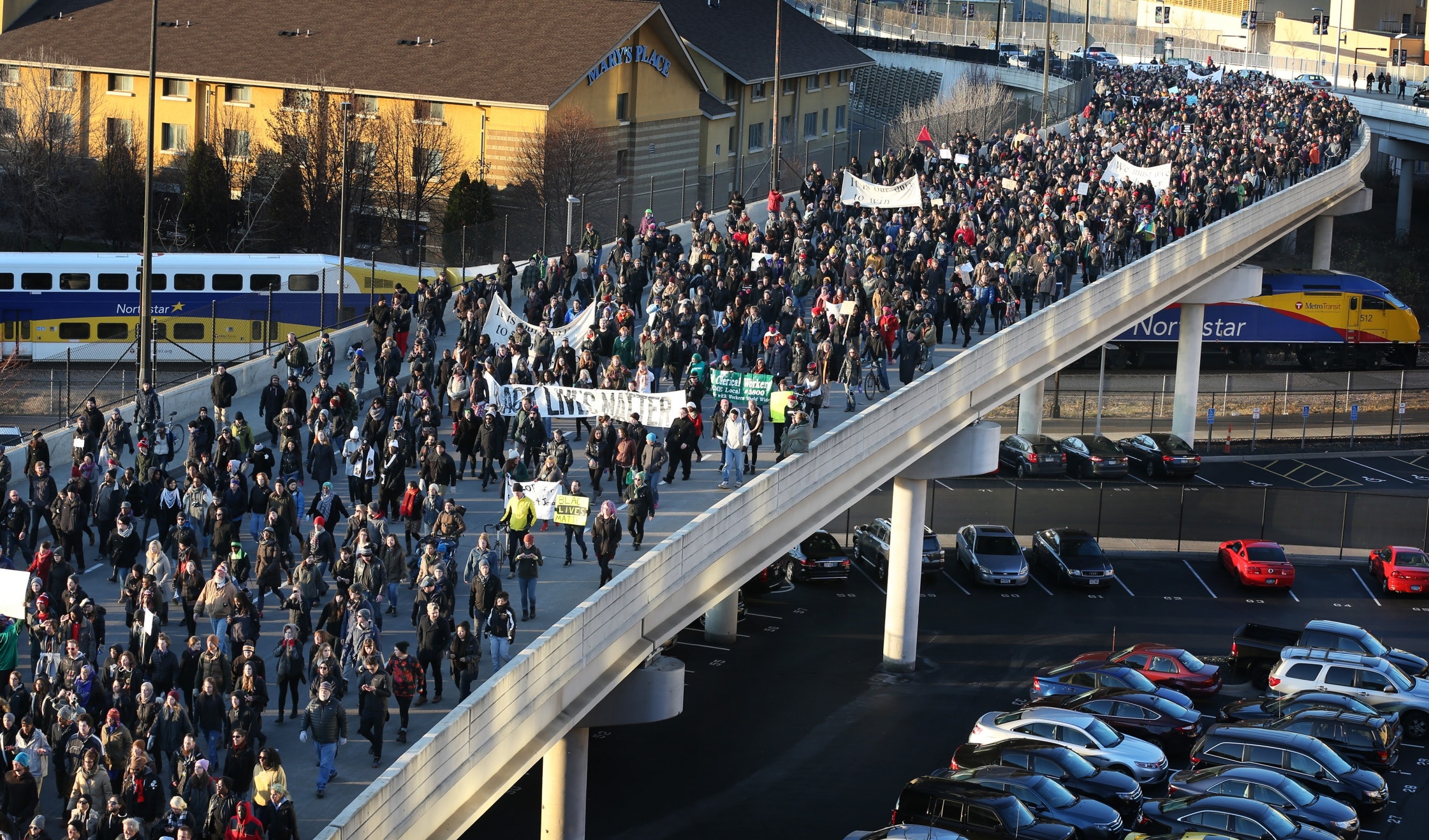 Hundreds of people are visible marching down a raised street, many holding signs 