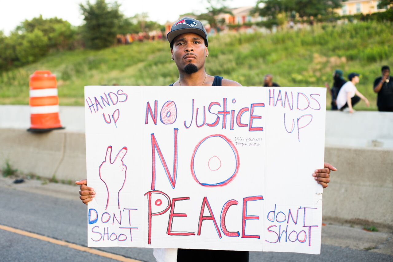 Protester stands with a sign reading, No Justice No Peace, Hands up, don't shoot.